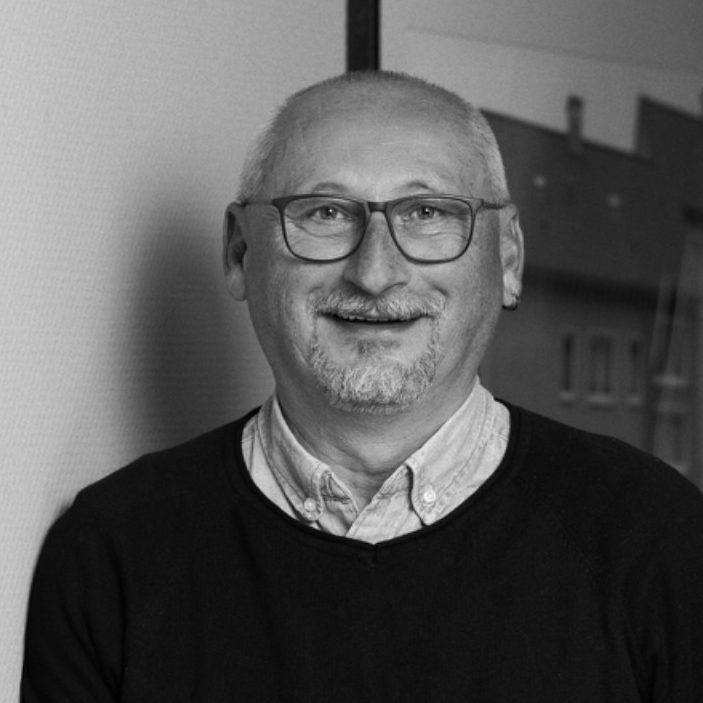 Ein lächelnder Mann mit Brille, in einem grauen Pullover vor einem Wandbild.