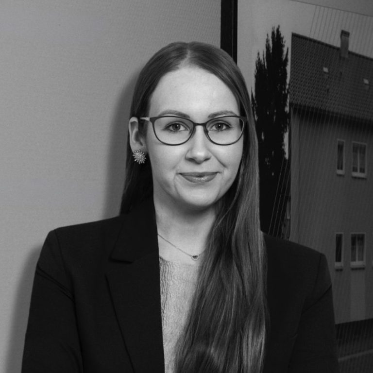 Eileen Ettner Junge Frau mit langen Haaren, Brille und blauer Bluse, steht vor einer Wand.