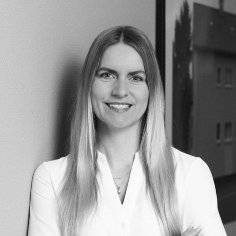 Blonde Frau mit langen Haaren lächelt, trägt ein weißes Hemd. Hintergründe sind unscharf.