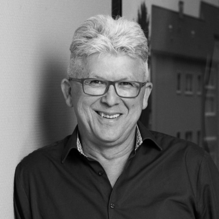 Lächelnder Mann mit Brille in einem dunklen Hemd vor neutralem Hintergrund.
