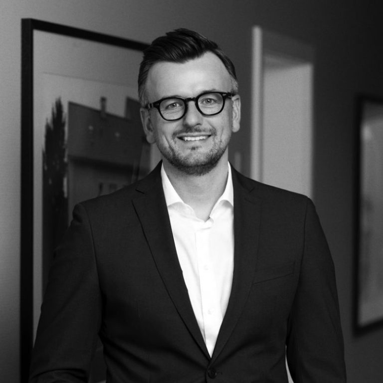Robin Zuhorn | Vorstand Männlicher Geschäftsmann in Anzug lächelt, sitzt am Tisch mit neutralem Hintergrund.