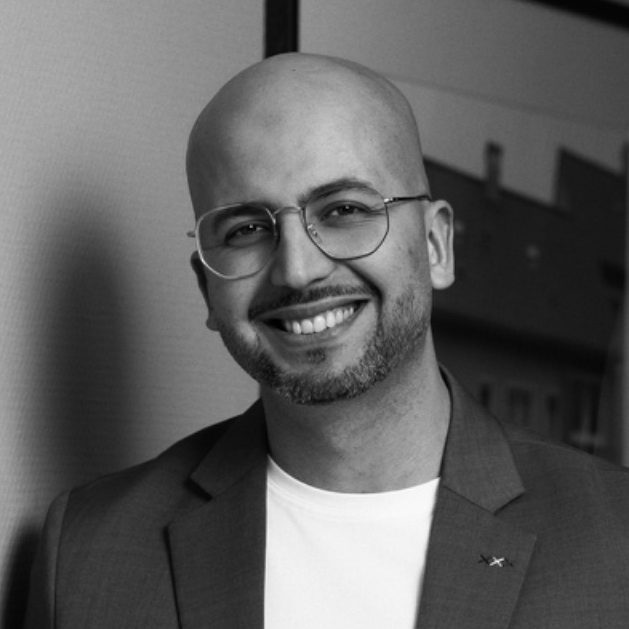 Mann mit Glatze und Brille, lächelt in einem grauen Blazer vor einem neutralen Hintergrund.