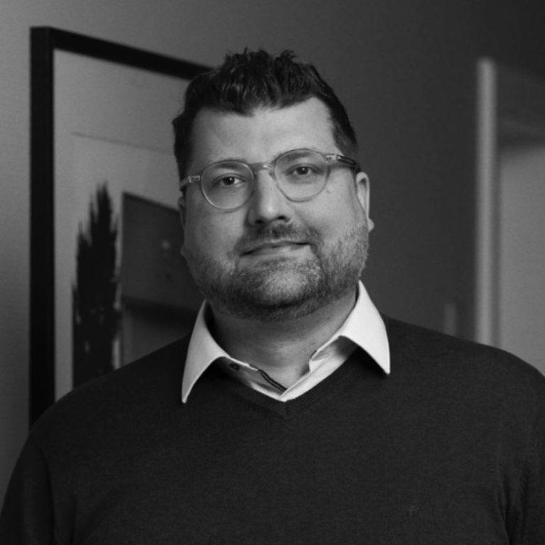 Porträt eines Mannes mit Brille und Pullover, lächelnd vor neutralem Hintergrund.