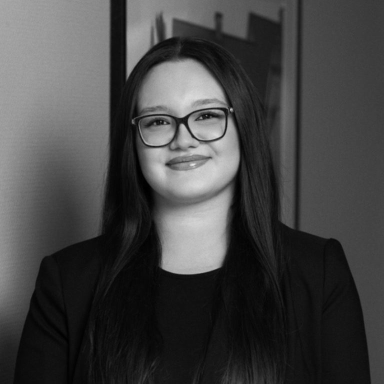 Azelya Kocagöz Junge Frau mit langen, dunklen Haaren und Brille in formeller Kleidung, lächelnd.