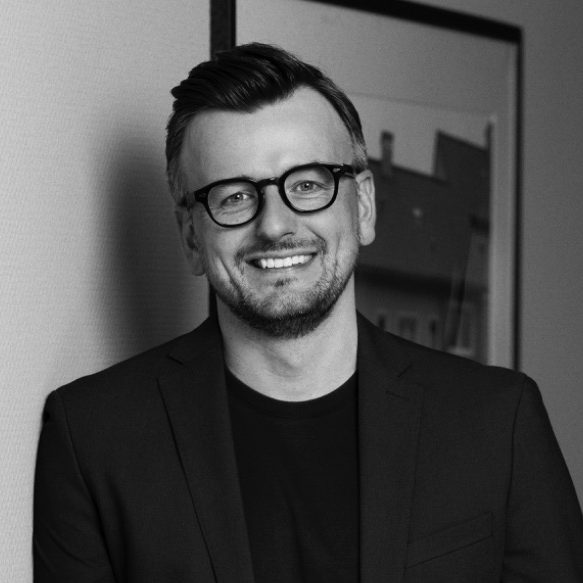 Mann mit Brille und schwarzem Blazer, lächelnd vor einem hellen Hintergrund.