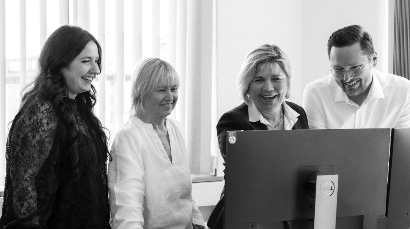 Unser Team der Handwerksbau AG Vier Personen stehen um einen Computerbildschirm und lächeln.