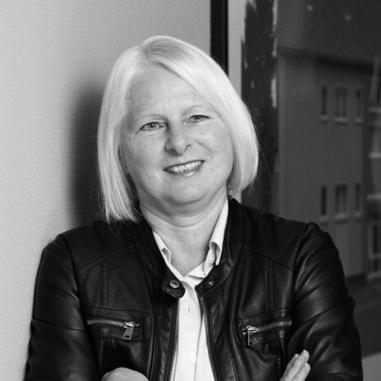 Blonde Frau mit lässiger Jacke lächelt, steht mit versetzten Armen vor einer Wand.