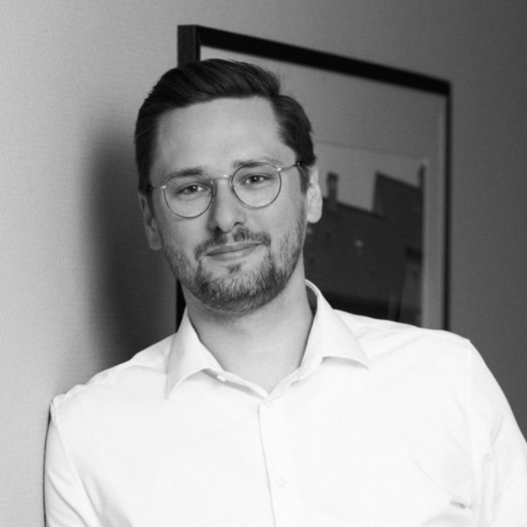 Ein lächelnder Mann mit Brille, in einem hellen Hemd, vor einer abstrakten Wandgestaltung.