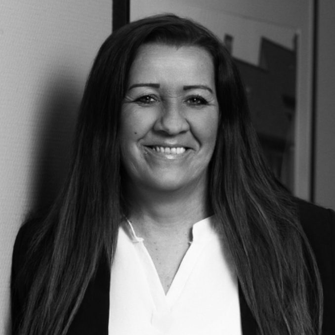 Eine Frau mit langen Haaren, lächelnd, trägt eine weiße Bluse und einen dunklen Blazer.